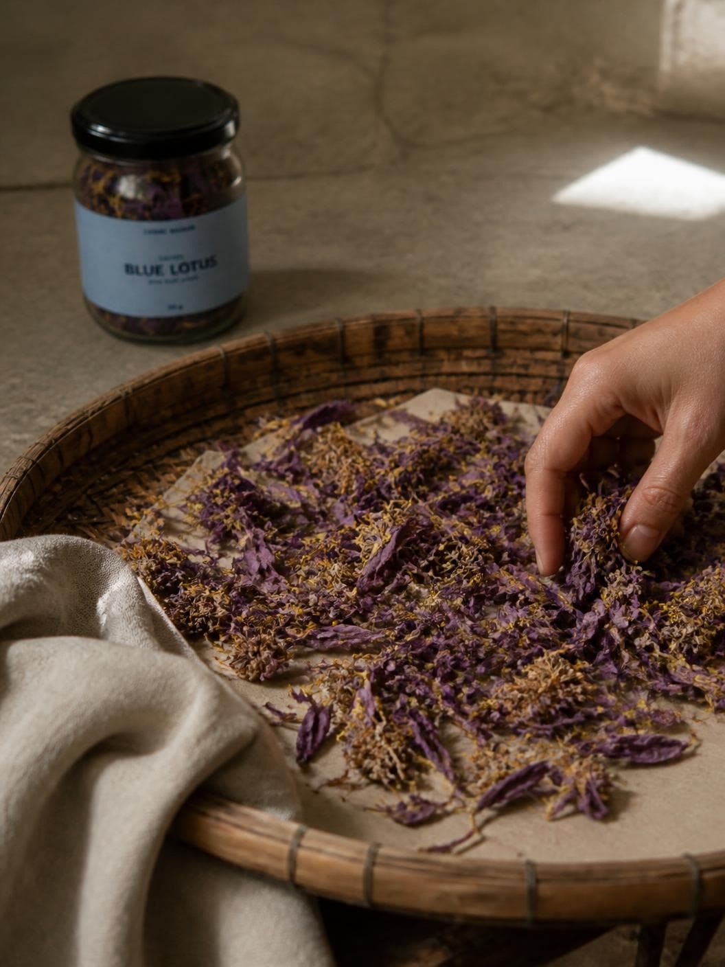 A wide woven drying tray on a stone floor holding a loose layer of dried violet petals and golden stamens, one hand turning them over, the jar on the floor behind.