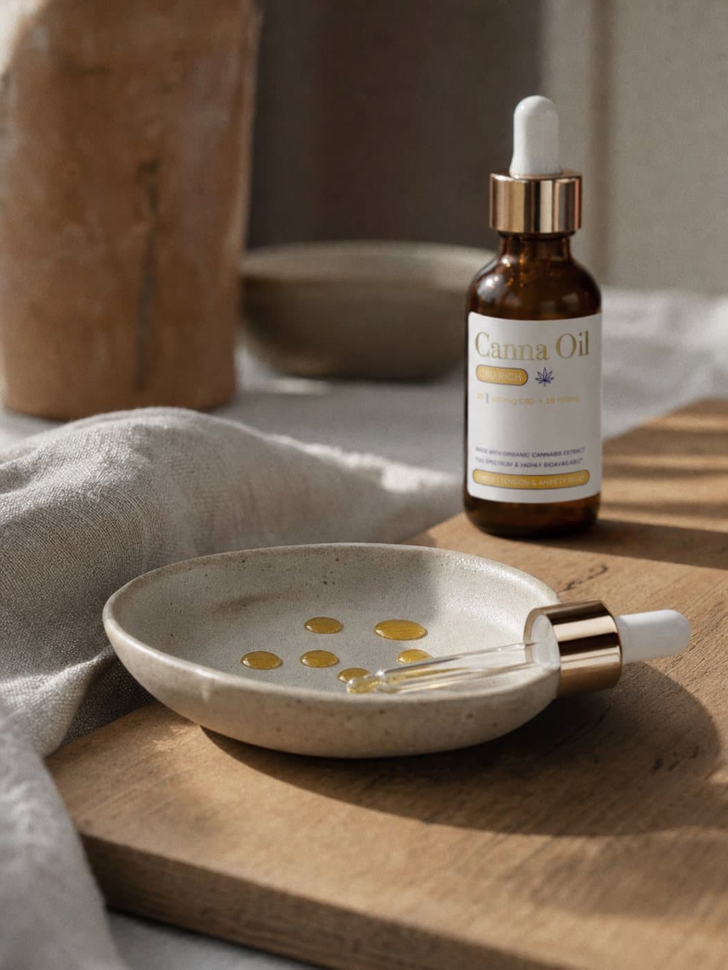 A shallow ceramic dish holding a handful of counted beads of amber oil, the glass pipette resting across its rim, the bottle standing behind and softly out of focus.