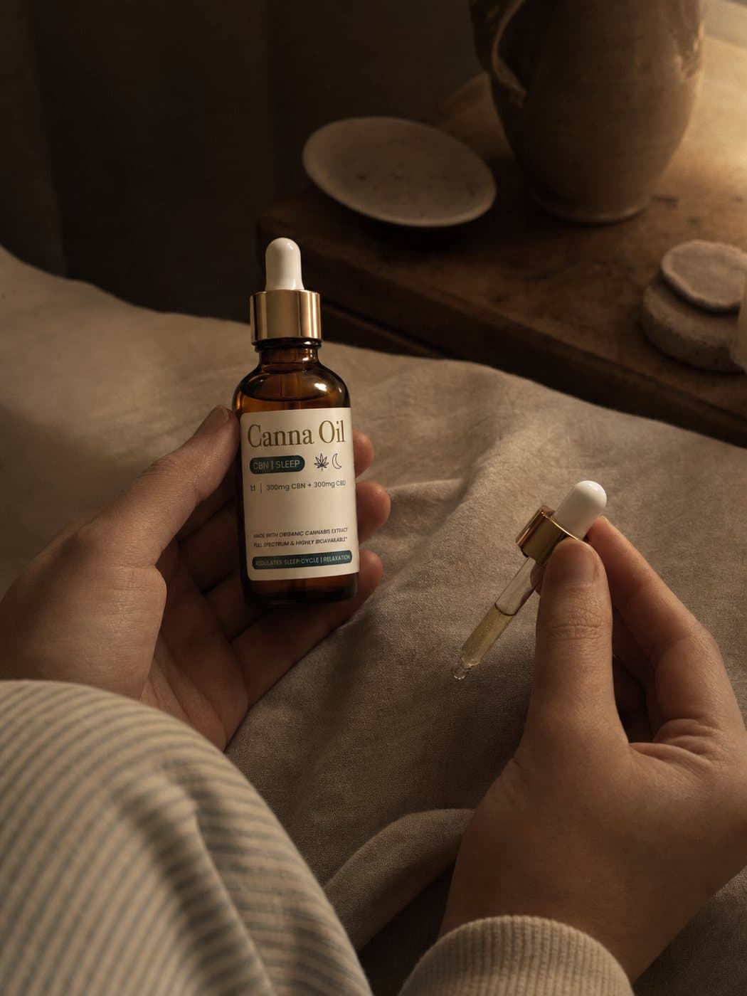 Two hands on a bed in lamplight, one holding the bottle and the other its glass pipette.