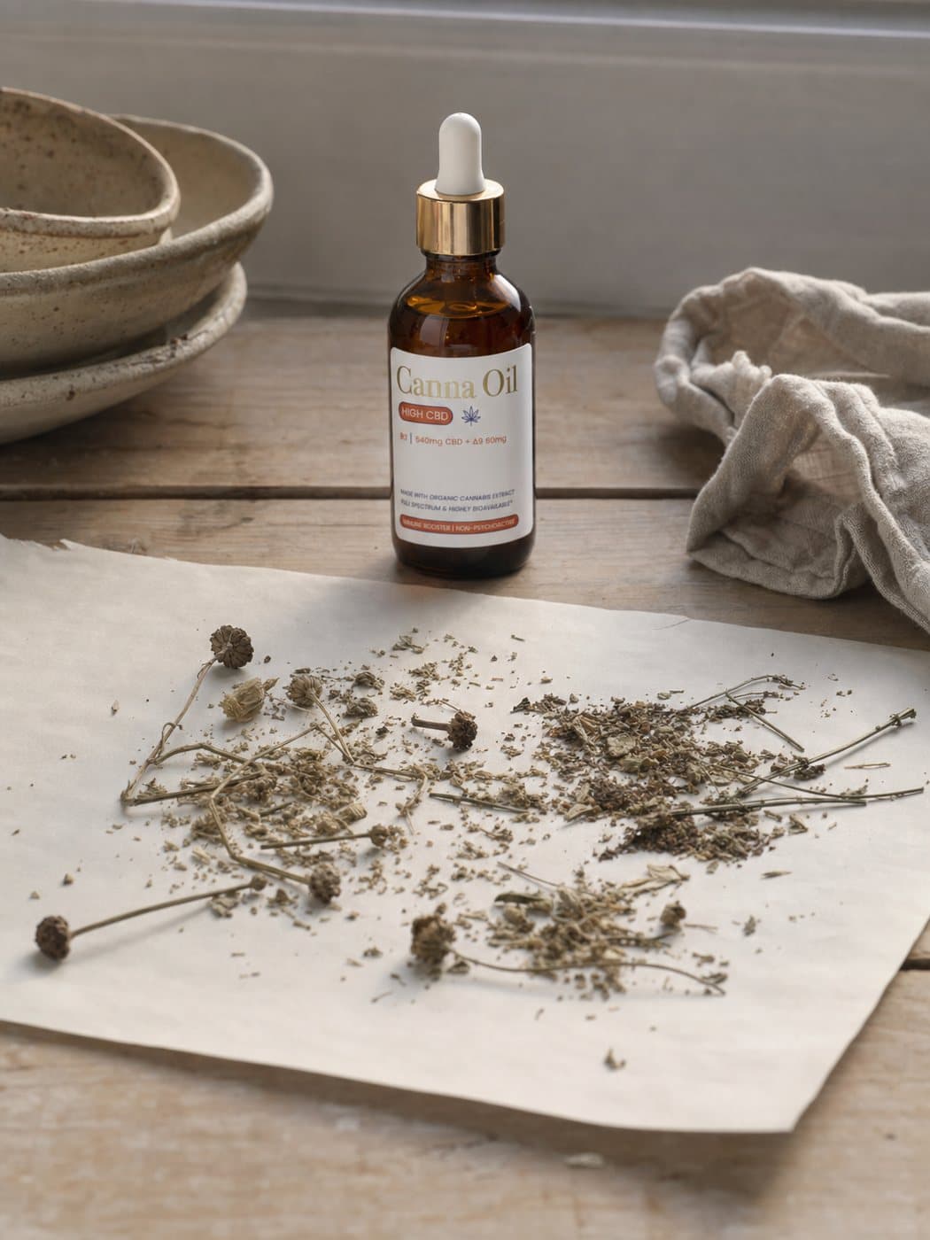 Dried whole botanical material — stems, seed heads and crumbled leaf — spread across a sheet of uncoated paper, the bottle standing behind it.