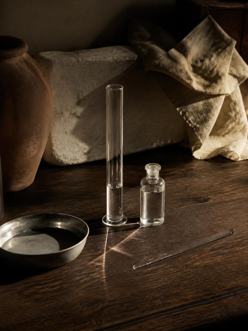A tall plain glass measuring cylinder holding a shallow depth of clear colourless liquid on a dark oiled walnut bench, a small unlabelled stoppered glass vial of the same beside it, low sun throwing a bright caustic streak across the timber between them, a shallow steel dish and a glass stirring rod nearby, a terracotta jar and a rough pale stone block behind.