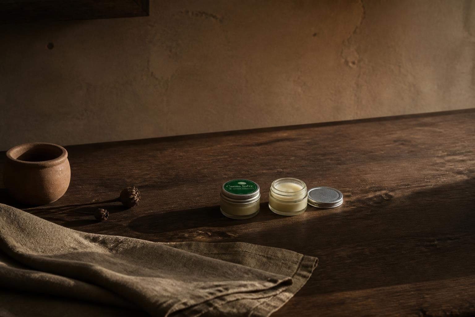 Two small glass jars of pale balm on a dark oiled walnut bench in low late-afternoon sun, the left one closed under a deep green lid label, the right one open with its silver lid beside it, long shadows running off across the timber, a squat terracotta pot and two dried seed heads at the left and heavy sand-coloured linen folded in front.