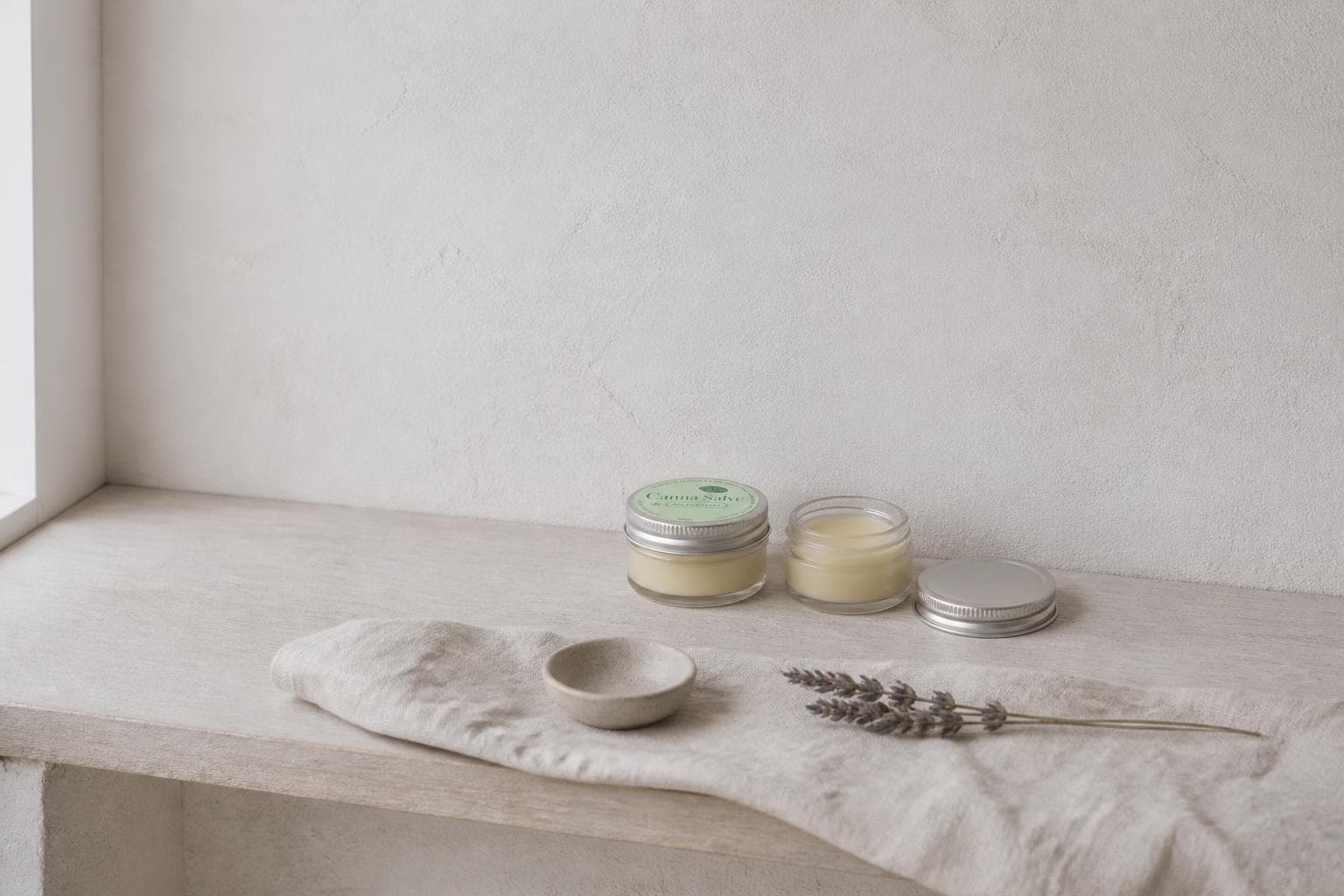 Two small glass jars of pale ivory balm on a whitewashed plaster shelf in cool morning light, the left one closed under a pale sage-green lid label, the right one open with its silver lid lying beside it, a folded bone-white linen cloth, a small stoneware dish and a sprig of dried lavender on the shelf in front.